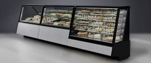 A large refrigerated display case filled with a variety of cakes and pastries, arranged on multiple shelves inside a glass enclosure with a modern, textured base.