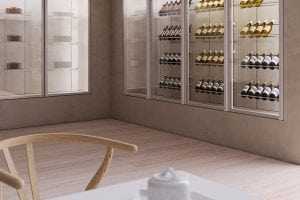 A modern wine cellar with glass-fronted shelves displaying bottles of wine. In the foreground, part of a wooden chair and a table with a white teapot are visible. The space has a minimalist, clean design.