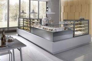 Modern bakery with large glass display cases filled with assorted pastries and desserts. The shop has large windows, white walls, and minimalist decor with pendant lights hanging from the ceiling.