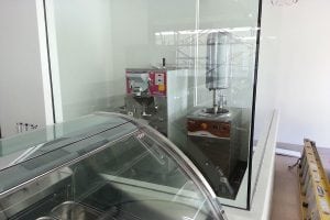 A modern gelato shop with commercial ice cream machines behind a glass partition, and a curved glass display case in the foreground. A yellow ladder is leaning against the wall on the right.