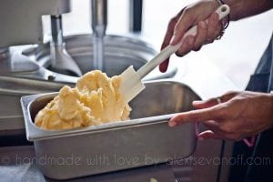 A person uses a white spatula to transfer creamy yellow gelato into a metal container in a kitchen, with a gelato machine visible in the background.
