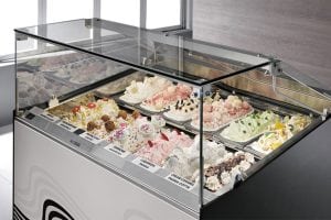 A glass display case filled with various colorful gelato flavors, each labeled with a name. The gelato is decorated with toppings, and the case is located in a well-lit, modern shop.