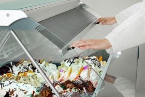 A person lifts the glass display cover of an ice cream or gelato case filled with various colorful flavors and toppings.