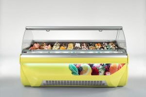 A yellow gelato display case features various colorful gelato flavors with toppings, visible through a clear glass cover. The case is decorated with images of fruit on the front.