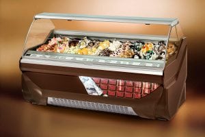 A display case filled with a variety of colorful gelato flavors, each topped with different decorations, in a well-lit, modern freezer with a glass front and brown exterior.
