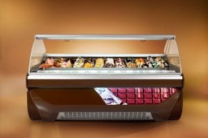 A gelato display case with a glass front, showcasing various colorful flavors of gelato arranged in rows, set against a warm brown background. The case features a decorative image of chocolate on the lower front panel.