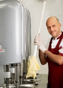 A person in a maroon apron and gloves uses a large spatula to handle creamy gelato from an industrial gelato-making machine in a kitchen.