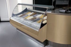 A refrigerated display case filled with various flavors of gelato, neatly arranged in rows. The case has a curved glass front and is part of a modern, beige and silver counter in a clean, bright setting.