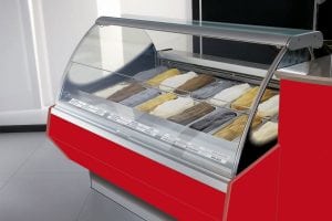 A red ice cream display case with a glass cover showing multiple trays of different colored gelato flavors, arranged in rows. Each tray is labeled, and the case is set in a clean, modern shop interior.