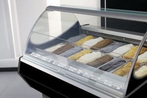 A glass display case filled with various flavors of gelato, arranged in metal trays. Each tray contains a different color and flavor, with labels in front of each section. The display is in a modern, clean setting.