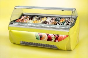 A yellow gelato display case filled with various colorful gelato flavors, topped with fruits and decorations, against a plain yellow background. The front of the case features images of assorted fruits.