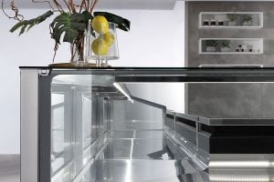 A modern, empty glass display case with a black frame sits in a minimalist kitchen. On top, a glass vase holds lemons and green leaves. Shelves with small jars are visible on a gray wall in the background.