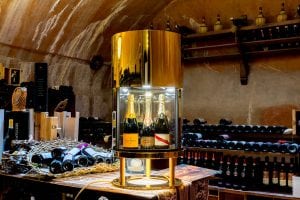 A gold cylindrical wine cooler displaying three bottles of champagne stands on a table surrounded by wine bottles, inside a rustic wine cellar with shelves of bottles and warm lighting.