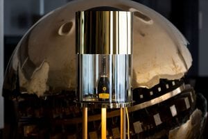 A bottle of Veuve Clicquot champagne is displayed inside a modern, cylindrical gold and glass case, positioned in front of a large, polished metallic sculpture with geometric cutouts.