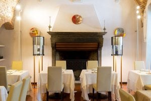 Elegant restaurant interior with cream-colored chairs, white tablecloths, and neatly set tables. A large stone fireplace is centered on the back wall, flanked by modern lamps, and decorated with contemporary art plates.