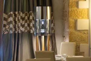 A modern dining area with cream chairs and a white table, set for a meal. A wine bottle is displayed in a sleek, cylindrical glass and metal case mounted on a pole near wavy-patterned curtains and a stone wall.