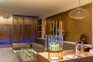 Modern lounge area with gold accents, a display of juice bottles, water bottles, snacks on a counter, gray cushioned seating, decorative lighting, and a partition with blue accent lighting in the background.
