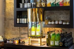 A gold and glass display case with green bottles stands on a counter in front of shelves stocked with oil bottles, jars, and packaged food items against a stone wall.