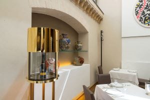 A modern dining room with set tables, a gold and glass wine display case, and decorative ceramic vases on glass shelves against a white wall. Abstract art hangs on the wall above.