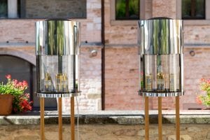 Two modern, cylindrical drink coolers with shiny metal tops and glass middles stand on wooden legs outdoors. Bottles and cans of beverages are visible inside. A stone building and potted flowers are in the background.