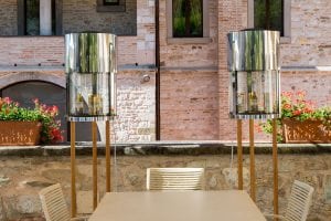 A modern outdoor dining area with a light-colored table and chairs. Behind the table are two tall, cylindrical glass and metal display cases containing bottles, set against a brick wall with flower boxes holding red geraniums.