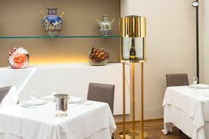 A modern restaurant interior with white tablecloths, set tables, a gold floor-standing wine cooler, and decorative ceramic vases displayed on shelves against a beige wall.