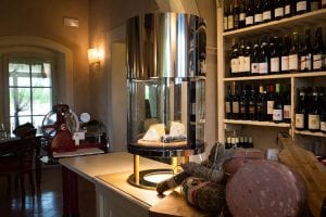 A cozy room with wine bottles on shelves, assorted cured meats and cheeses on a wooden counter, and a glass cheese display case in the center. A red meat slicer stands near a window letting in natural light.