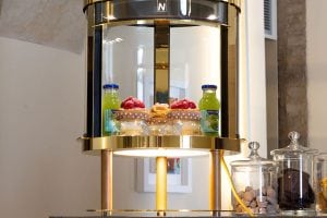 A display case with gold and glass accents showcasing pastries, two bottles of green juice, and jars of sugar cubes and chocolates on a counter in a well-lit room.