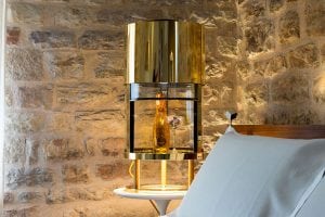 A gold and glass champagne cooler with a bottle and glasses inside sits on a small white table next to a stone wall and a bed with white bedding.