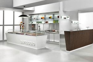 Modern, minimalist chocolate shop interior with a white display counter filled with assorted chocolates, shelves with decorative items, and a wooden side counter against clean white walls and floor.