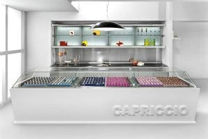 A modern white display counter labeled CAPRICCIO showcases assorted chocolates in neat rows; behind it are glass shelves with decorative items and wine glasses in a stylish, minimalist shop interior.
