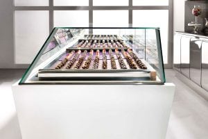 A modern glass display case filled with neatly arranged rows of assorted chocolates or pastries in a bright, minimalist shop setting.