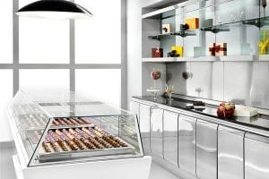 A modern, sleek chocolate shop with a glass display case filled with assorted chocolates. Shelves behind the counter hold decorative gift boxes and glass jars with candies. The room features stainless steel surfaces and minimalist decor.