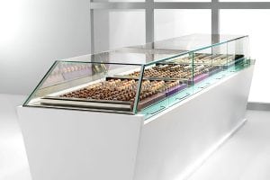 A modern glass display case filled with assorted chocolates and pastries is shown in a bright, minimalistic setting with white walls and large windows in the background.