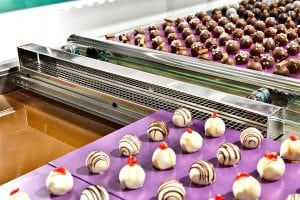 A factory conveyor with rows of decorated chocolate truffles, both dark and white, on purple trays, moving beside a machine filled with melted chocolate.