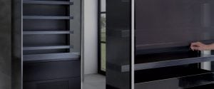 Two modern, empty, black display refrigerators stand next to each other indoors. One unit has open shelves; the other has a hand pulling open a tinted glass door. A window is visible in the background.