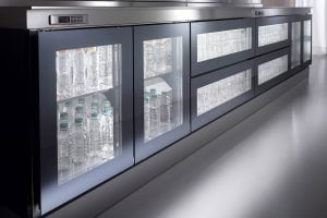 A modern commercial refrigerator with glass doors displays rows of clear plastic water bottles neatly arranged on shelves inside, with bright interior lighting.