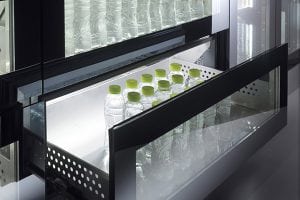 A partially open refrigerator drawer reveals several clear plastic water bottles with green caps neatly arranged inside, while other bottles are visible in the shelf above.