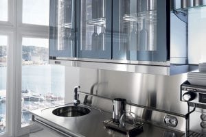 A modern stainless steel kitchen with a round sink, glass-front cabinets, a coffee machine, and a window overlooking a marina with boats and water outside.