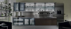 A modern kitchen with glass-front cabinets, open shelves holding bottles and bowls, and stainless steel countertops. Large windows on the left let in natural light, and potted plants line the window sills.