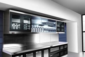 Modern kitchen with glass-fronted cabinets displaying neatly arranged glassware and dishes, a stainless steel countertop, and sleek black cabinetry below, set against a blue and white wall.
