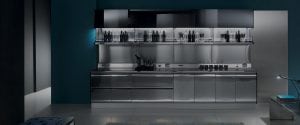 A modern kitchen with sleek, reflective stainless steel cabinets and appliances, open shelves holding bottles and glasses, under-cabinet lighting, and a dark blue accent wall.