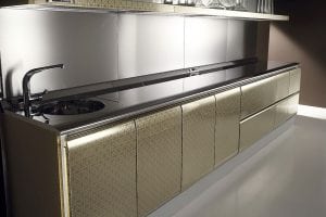 A modern kitchen with glossy gold-patterned cabinets, a sleek stainless steel countertop, an integrated round sink, and glassware stored on an upper shelf against a reflective metallic backsplash.