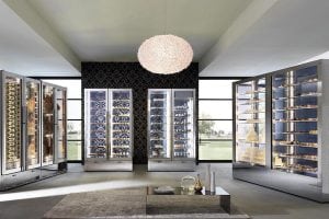 A modern, bright room with large glass display refrigerators filled with wine bottles, cheeses, and meats, set against a dark accent wall and decorated with a unique chandelier and minimalistic furnishings.
