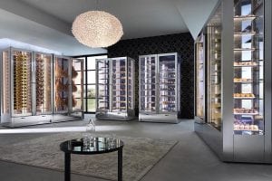 A modern, elegant room with large glass display refrigerators featuring pastries and cured meats. A round table and rug are in the center, and a unique chandelier hangs from the ceiling. Natural light streams in through tall windows.