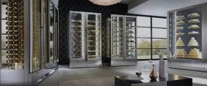 Modern showroom with tall glass display cases holding cakes, pastries, and bottled drinks, large windows letting in natural light, and contemporary decor with a patterned wall and a grey rug on the floor.