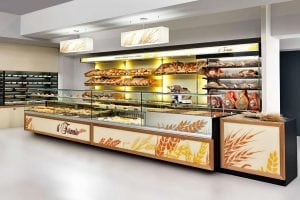 A modern bakery display with shelves of various breads, pastries, and meats behind glass counters. The counter has wheat designs, and the background is well-lit with a clean, organized appearance.
