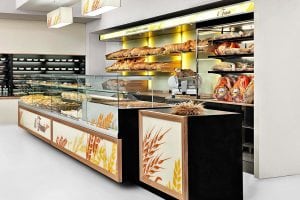 A modern bakery with glass display cases filled with pastries and bread. Shelves behind the counter display baguettes, loaves, and cured meats. The décor features wheat illustrations and warm lighting.