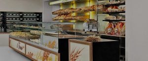 A modern bakery interior with glass display cases filled with pastries and bread, shelves of baguettes and loaves, and cured meats hanging on the right. The counter features wheat-themed decor.
