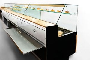 A modern bakery display case with glass panels, wooden accents, metal drawers, and shelves showcasing various pastries on trays. The counter is clean and minimalistic.
